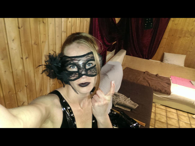 HeisseLina-hot with black lace mask, dark lipstick, and black shiny dress, flashing a middle finger, showing her pale tits in a wooden room with a bed and red curtains.