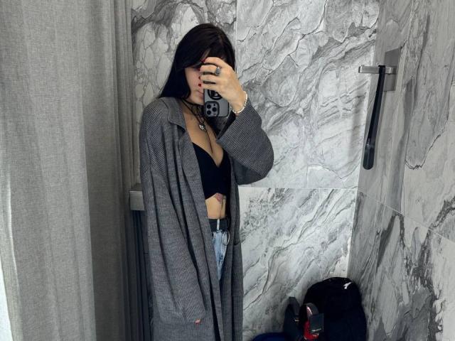 Tsukerberg with dark hair, wearing a grey coat, black bra showing her tits, holding a phone, taking a mirror selfie in a marble-tiled bathroom.