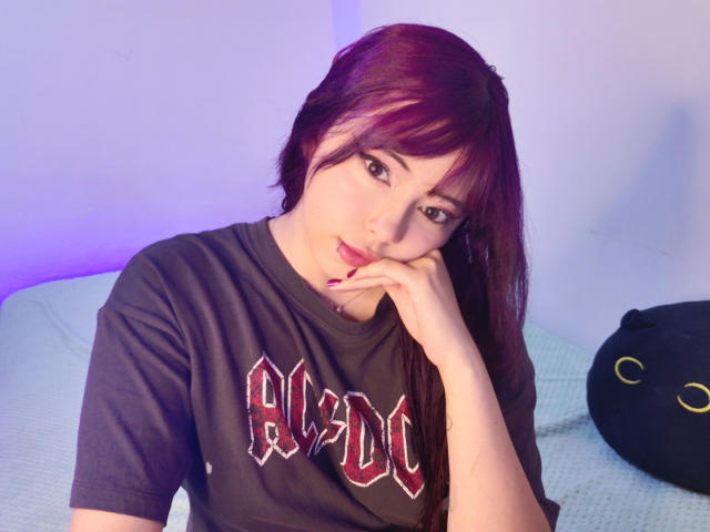 Arieyaara with red-purple hair, brown eyes, and fair skin, wearing a black "AC/DC" shirt, resting her chin on her hand, looking at the camera, sitting on a bed.