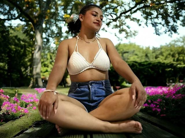 Ameliebx, avec une poitrine de taille moyenne, portant un haut de bikini en crochet blanc, assise en tailleur sur un banc en bois, vêtue de shorts en jean taille haute, dans un jardin ensoleillé avec des fleurs roses..