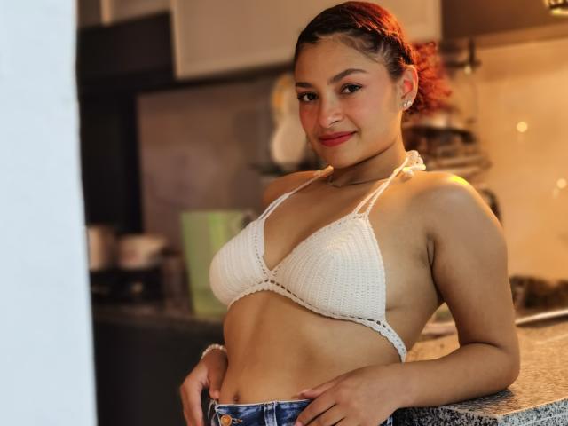 Ameliebx, con cabello rojo, de busto medio, con un top de bikini blanco de crochet, sonriendo en una cocina, vestida con jeans azules, de pie frente a una encimera de granito..