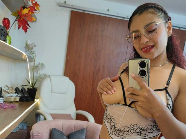 Ameliebx met een bril, rode lippenstift en gevlochten haar, die een selfie maakt in een spiegel, waarbij haar borsten te zien zijn in een zwart en roze kanten beha, en ze een gouden iPhone vasthoudt..
