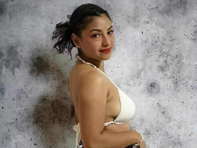 Ameliebx, com cabelo preto ondulado, usando um top de biquíni branco de crochê, mostrando os seios, sorrindo, com as mãos no bumbum, em pé contra um fundo cinza texturizado..