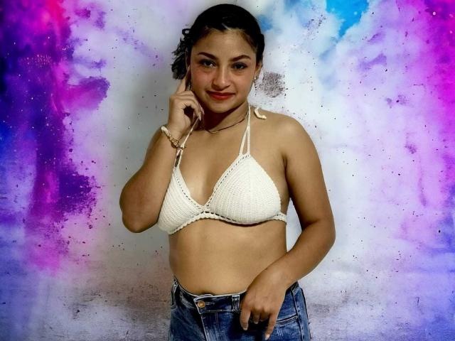 Ameliebx mit lockigem, schwarzem Haar, gebräunter Haut, trägt ein weißes, gehäkeltes Bikini-Oberteil, das ihre Brüste zeigt, und blaue Jeans, lächelt, berührt ihr Kinn, vor einem farbenfrohen Hintergrund in Lila und Blau..