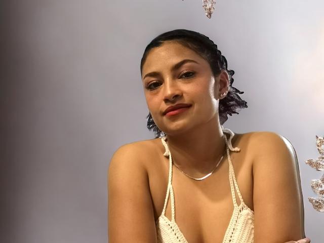Ameliebx, avec ses cheveux foncés tressés, porte un haut en crochet qui dévoile sa petite poitrine et affiche un sourire discret. Elle a la peau claire et porte un collier fin..