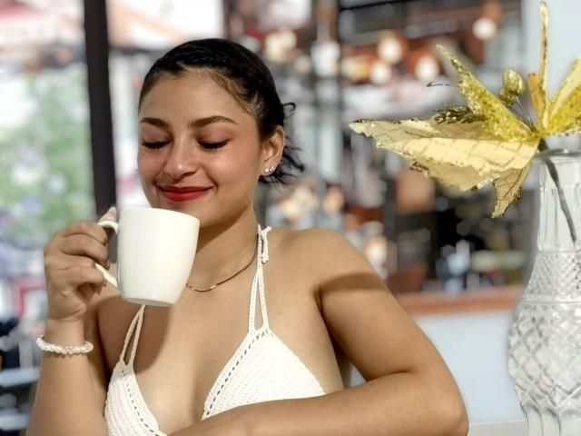 Ameliebx mit gebräunter Haut, schwarzen Zöpfen, kleinen Brüsten in einem weißen, gehäkelten Bikini-Oberteil, hält eine weiße Tasse, lächelt, trägt roten Lippenstift, ein weißes Armband, eine gelbe Blume in einer Glasvase..