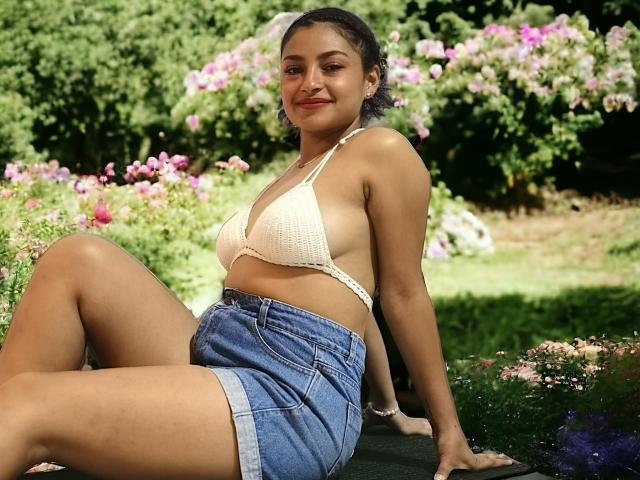 Ameliebx met een bruine huid, krullend donker haar en kleine borsten, draagt een witte bikini-top van kant en zit in blauwe denim shorts. Ze glimlacht in een tuin met roze bloemen..