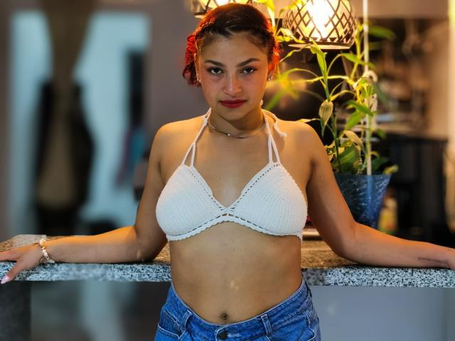 Ameliebx, avec ses cheveux roux bouclés, un haut de bikini en crochet blanc qui révèle ses petites seins, et un jean bleu, appuyée sur un comptoir en granit, regardant l'appareil photo avec un léger sourire..