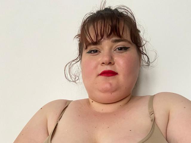 SarahSteelX ha un viso paffuto con rossetto rosso, capelli castani disordinati, eyeliner nero e una canotta beige, che mette in mostra il suo seno grande e la sua pelle morbida e chiara..