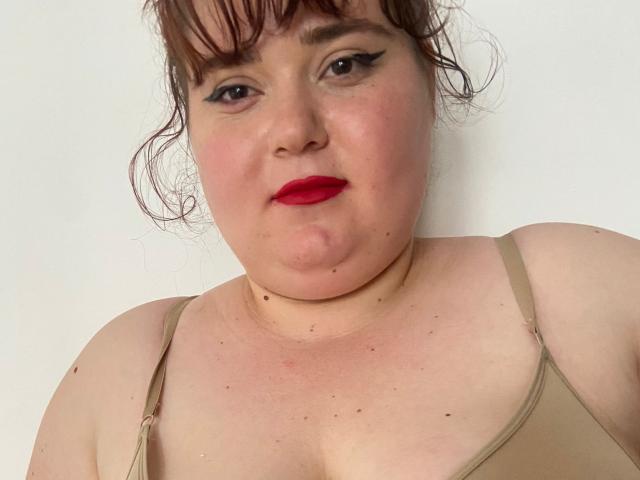 SarahSteelX com batom vermelho, delineador preto e cabelo castanho em um coque bagunçado, vestindo uma regata bege, mostrando seu rosto gordinho, peito e pequenas manchas..