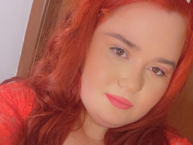 A SarahSteelX tem cabelo ruivo, sombra rosa nos olhos e batom vermelho. Ela está olhando para a câmera com um leve sorriso, vestindo uma blusa vermelha. Seus seios estão parcialmente visíveis..