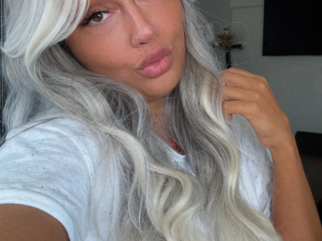EmmaFarrah69 with long, wavy, silver hair, pouty pink lips, and a white tee, holding her hair. Her tan skin and brown eyes are prominent.