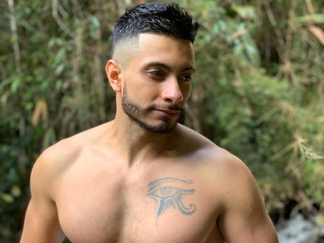 Shirtless MarkBilly69 with a tattoo of an eye on his right chest, short black hair, and a trimmed beard, looking down with a slight smile, in a forest.
