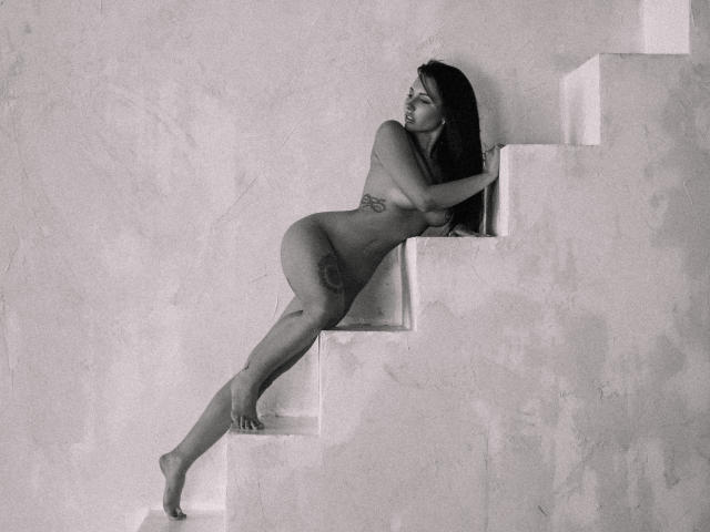 Fotografia in bianco e nero di una donna nuda, con tatuaggi, appoggiata a delle scale bianche, con le gambe distese e il sedere in alto, che guarda di lato. Ha il seno piccolo e lunghi capelli scuri..