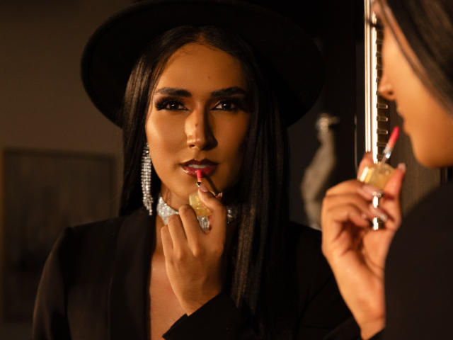 FallonRogers mit langem, schwarzem Haar, die einen schwarzen Hut und eine Jacke trägt, trägt roten Lippenstift auf ihre vollen Lippen mit einem Pinsel mit rosa Spitze auf und blickt in einen Spiegel..