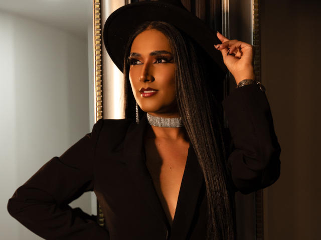 FallonRogers with long black braids, wearing a black hat, choker, and blazer, looking at camera with hand on hat, sexy brown skin, glossy pink lips, stylish and confident.
