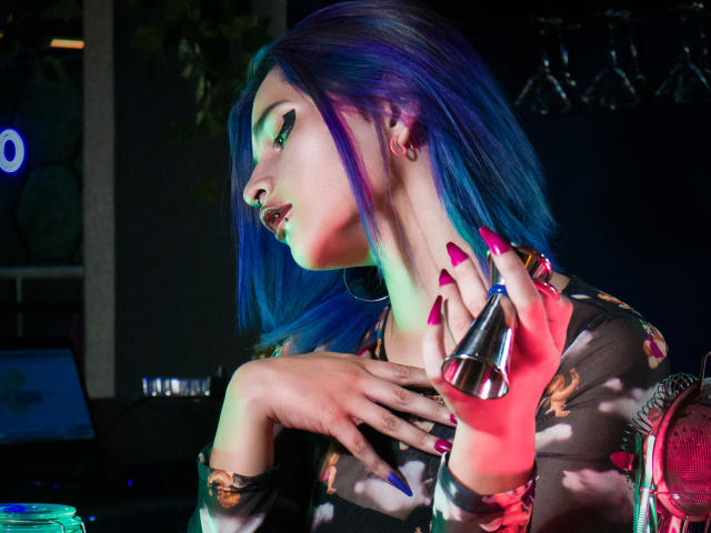 MaiaThunder con riflessi blu, indossa una maglia scura con motivi floreali, tiene un anello metallico, ha unghie lunghe rosa, orecchini a cerchio, guarda di lato, illuminata da luci colorate, in un ambiente da discoteca..