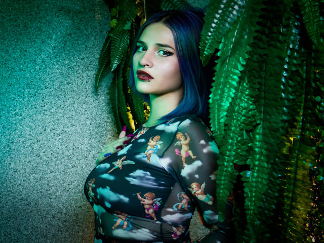 MaiaThunder with blue hair, wearing a sheer top with fairy patterns, has large tits and a serious expression, standing amid green ferns with green and blue lighting.