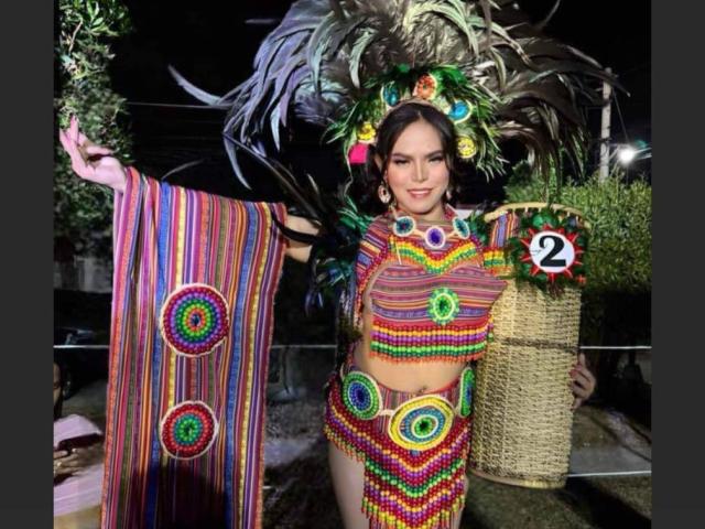 AnastasiaPoks with colorful, striped traditional outfit featuring large circular patterns, holding a woven basket, wearing a feathered headdress, and a number 2 pin, in a nighttime setting.