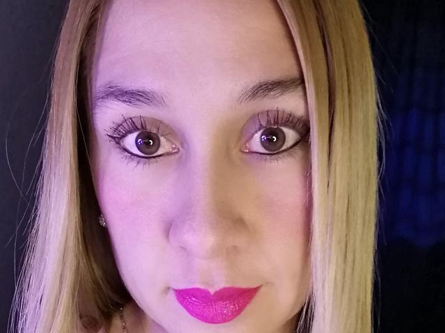 Dana69s has blonde hair, brown eyes, thick eyelashes, and bright pink lipstick. Her skin is fair, and she's looking directly at the camera with a slight smile.