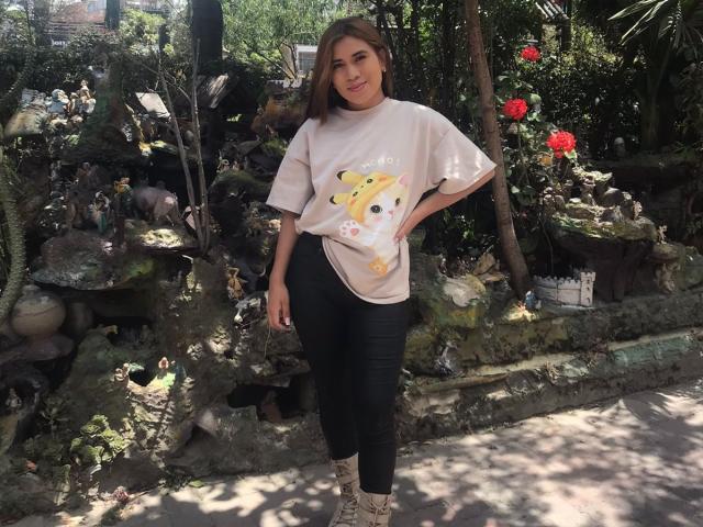 BellaBickers, medium-ass, wearing a loose white shirt with a yellow dog graphic, black tight pants, and snakeskin boots, standing in a sunny, rock-filled garden.