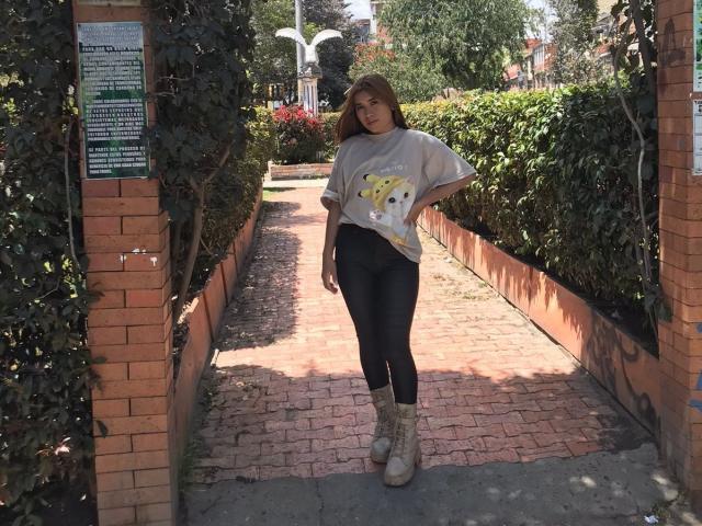 Bella, avec ses cheveux bruns, portant une chemise grise avec un chat jaune, un legging noir et des bottes beige, se tient sur un chemin en briques entre des murs en briques et des murs verts..