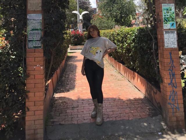 BellaBickers stands in a brick walkway, wearing a loose white shirt with a yellow graphic, black leggings, and beige boots, with a relaxed smile and hands on her hip.