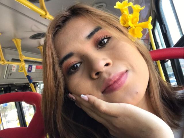 Bella, cheveux bruns, fleur jaune dans les cheveux, siège de bus rouge, barres de maintien jaunes, souriante, lèvres roses, main sous le menton, ongles roses, maquillage, yeux marrons, intérieur de bus..