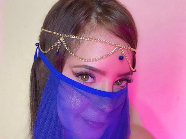 HoneyFoxy has brown eyes, wearing a gold chain headpiece with blue jewels, and a blue sheer mask covering her mouth, with pink and white lighting.