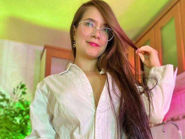 HoneyFoxy with long brown hair, glasses, pink lipstick, white button-up shirt, holding hair, smiling, green and pink light in background, wooden cabinets.