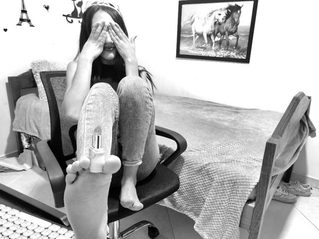 KiriamGomez sentada em uma cadeira, cobrindo o rosto com as mãos, vestindo jeans e sem sapatos, mostrando seu pé descalço com um pequeno objeto preso à sola..