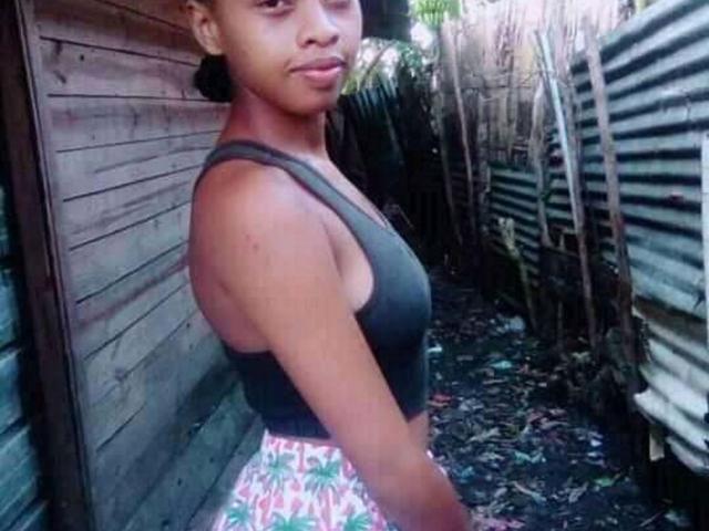 ISmileLove has dark brown skin, small tits, and wears a green tank top with pink floral pants, standing in a narrow, wooden, and corrugated metal-sided alleyway.