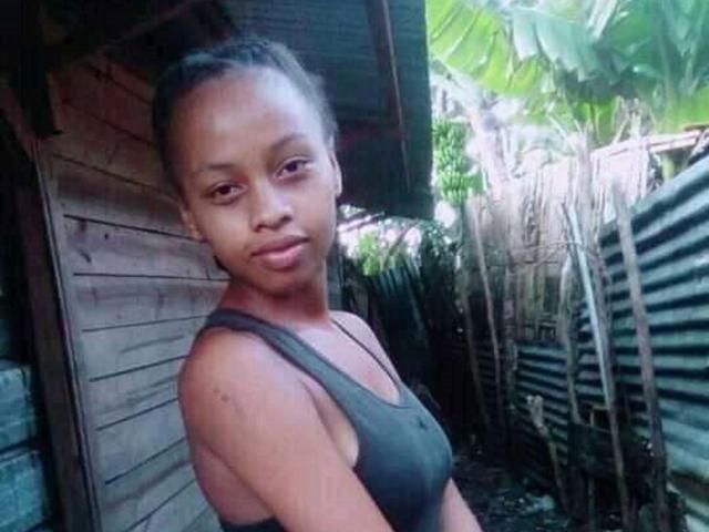 ISmileLove has dark skin, small tits, and a slim body. She's wearing a gray tank top, looking at the camera with a slight smile, and has her hair in a bun.