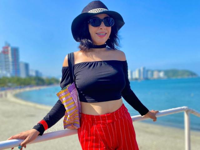 AnastasiaEvansX in a black off-shoulder top, red pinstriped pants, black hat, sunglasses, and choker, holding a colorful bag, standing by a beach railing, smiling.