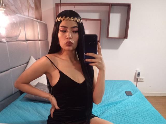 MelodyBellanova with long black hair, wearing a black velvet dress and gold headpiece, pouting at the camera, holding a phone, sitting on a blue bed. Boobs visible.