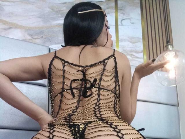 MelodyBellanova with black hair, wearing a gold chain mesh top, exposing her back and butt, holding a light bulb, sitting on a white couch.