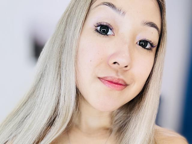 RenyLime's face, featuring blonde hair, blue eyes, long lashes, pink lips, and a slight smile, with a soft, fair complexion and a small beauty mark on her right cheek.