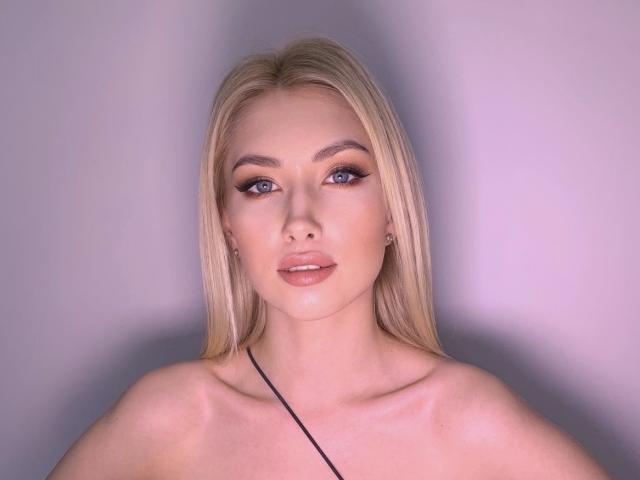 MashaTitova has blonde hair, blue eyes, and full lips with subtle pink lipstick. She's wearing a black strap and has a slight sheen on her face.