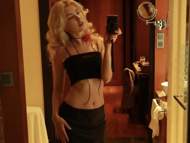 MashaTitova with blonde hair, wearing a black strapless top and skirt, showing a slim midriff, holding a phone, and looking at the mirror with a slightly open mouth.