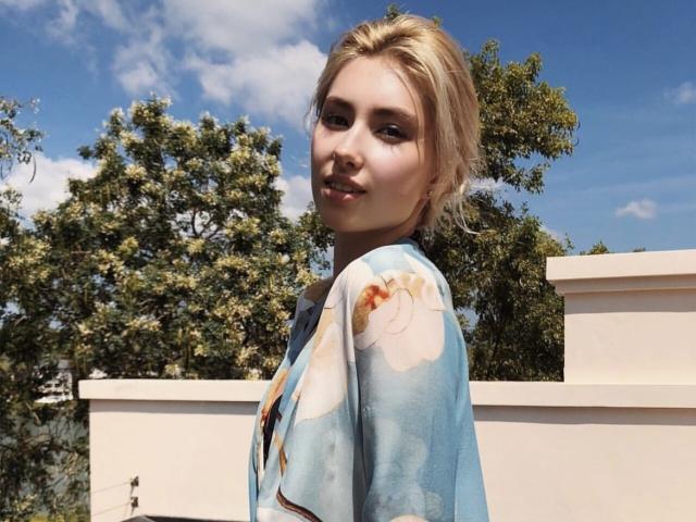 MashaTitova with blonde hair, wearing a blue floral kimono, standing outside, looking over her shoulder, clear blue sky, fluffy white clouds, and green trees in the background.