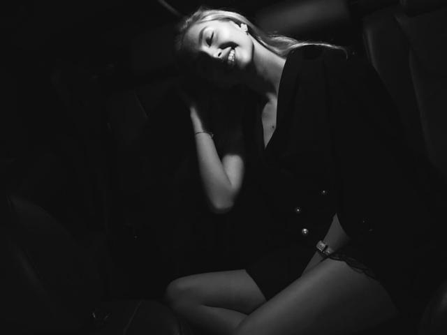 MashaTitova in a black dress, sitting in a dark car, smiling with eyes closed, hand on head, exposed thighs, long blonde hair, dimly lit, black and white.