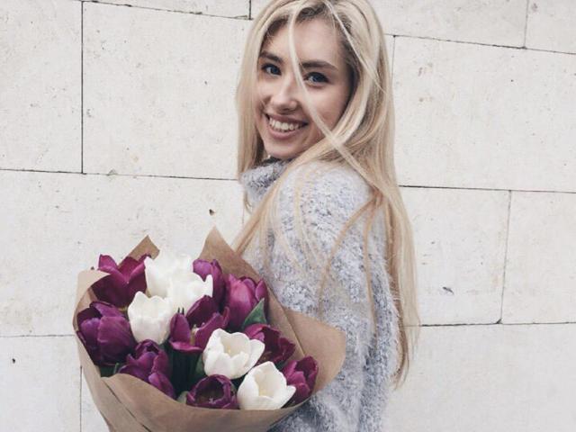 MashaTitova with long blonde hair, smiling, wearing a gray fuzzy sweater, holding a bouquet of purple and white tulips. Boobs not visible. Ass not visible. Pussy not visible.