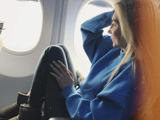 MashaTitova with long blonde hair, blue hoodie, and dark pants, sitting with knees up, hand in hair, looking out airplane window, smiling softly.