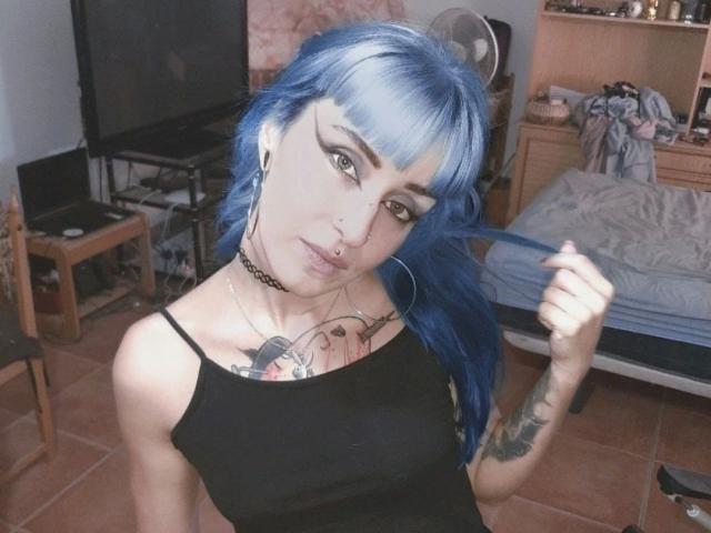 HannaDown-hot with blue hair, black tank top, tattoos, nose ring, choker, and hoop earrings, holding hair, showing tits, in a messy room, with a bed in the background.