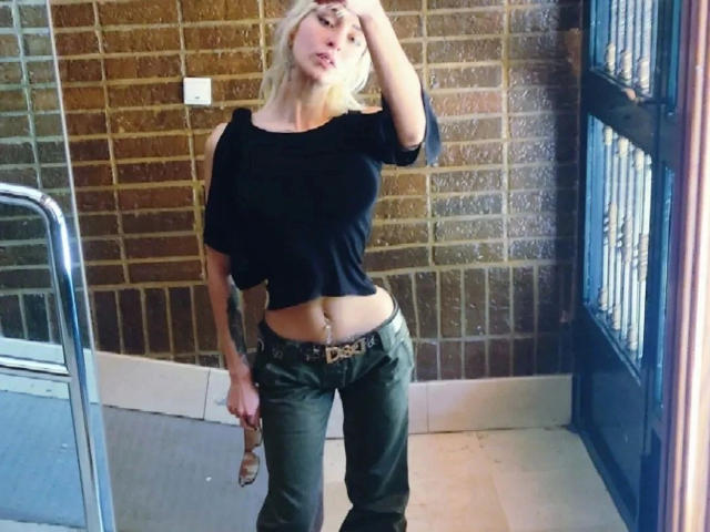 HannaDown, met blond haar, een zwarte top, groene broek en een zichtbare navel, houdt een zonnebril vast, staat in een kamer met bakstenen muren en kijkt naar de camera met een hand op haar hoofd..