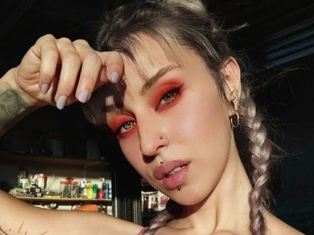 Hanna, met paarse gevlekte vlechten, rode oogschaduw, een neusring, een lip piercing en een getatoeëerde arm, heft haar hand op, waarbij haar glanzende, roze lippen en haar donkere, door de zon verlichte gezicht te zien zijn..