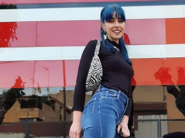 Hanna, con el pelo azul, lleva una camiseta negra y pantalones vaqueros de cintura alta, un bolso con estampado de cebra, está sonriendo, mostrando ligeramente su trasero, y está de pie frente a una pared a rayas rosa y blanca..