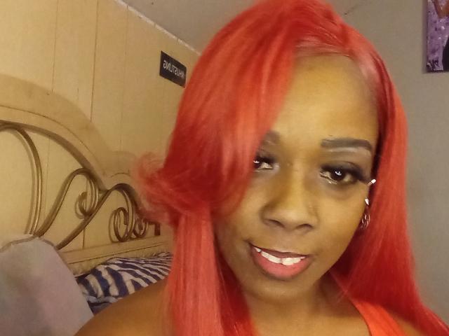 JuiceTiax with red hair, dark skin, and small nose, wearing a red top, smiling with pink lips, and a nose ring on the right side. Bedroom setting.