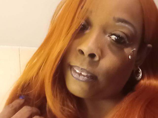 JuiceTiax with shiny orange hair, silver glitter lips, hoop earrings, and a small facial scar, looking slightly to the side with a subtle smile.