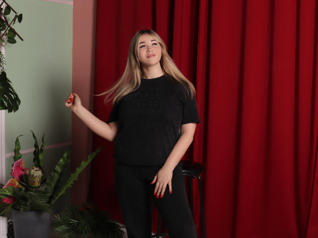 SarahStar, avec de longs cheveux blonds, un t-shirt noir, un pantalon noir et des ongles rouges, debout, la main droite levée, la main gauche sur sa cuisse, un léger sourire, rideaux rouges, mur vert..